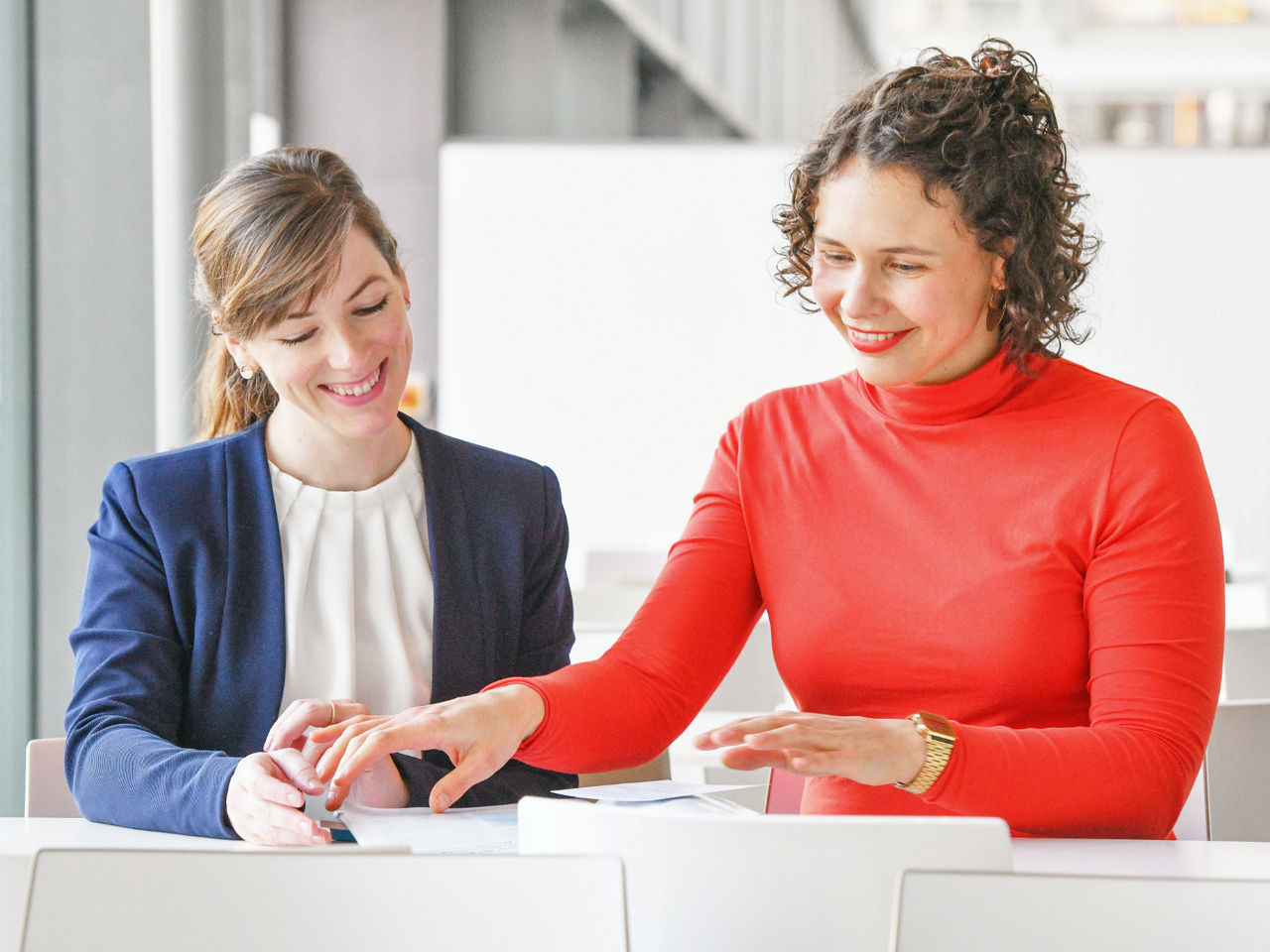 Zwei Frauen sitzen an einem Tisch in einem modernen, hellen Raum und lächeln, während sie gemeinsam auf ein Dokument oder Tablet schauen. Eine der Frauen zeigt dabei gestikulierend auf das Gerät.