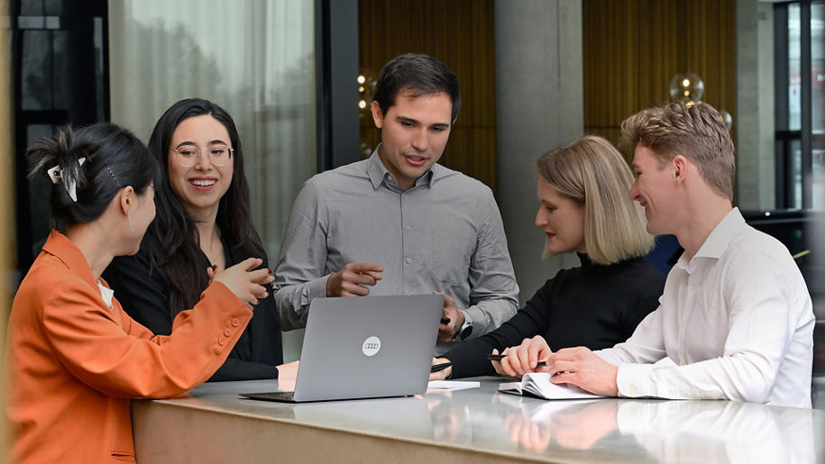 Fünf Personen stehen an einem Tisch mit einem Laptop in der Mitte und unterhalten sich
