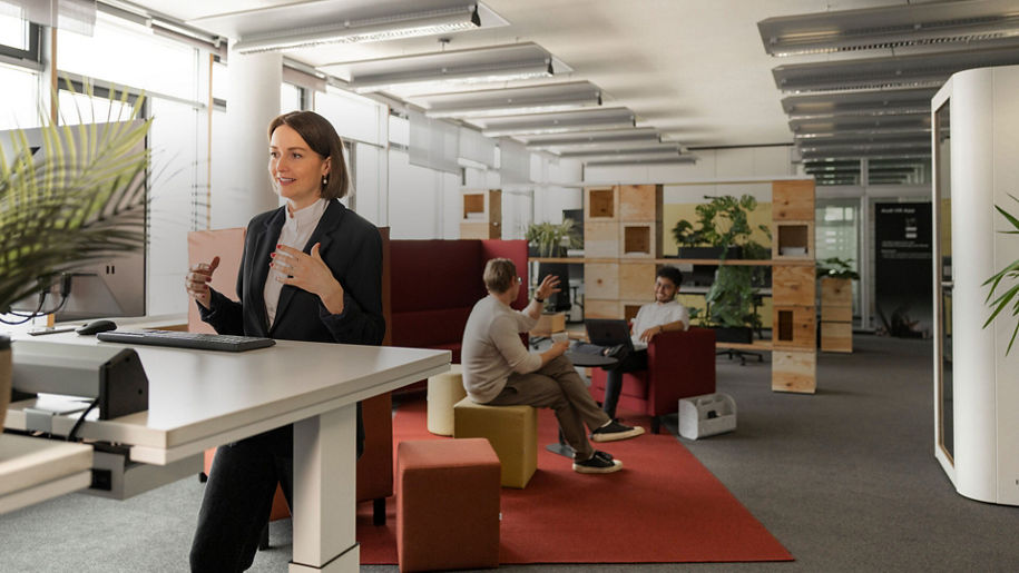 Drei Personen befinden sich in einem Büro, zwei sitzen und unterhalten sich und eine Frau steht an einem Schreibtisch und spricht in einen Bildschirm eines Computers