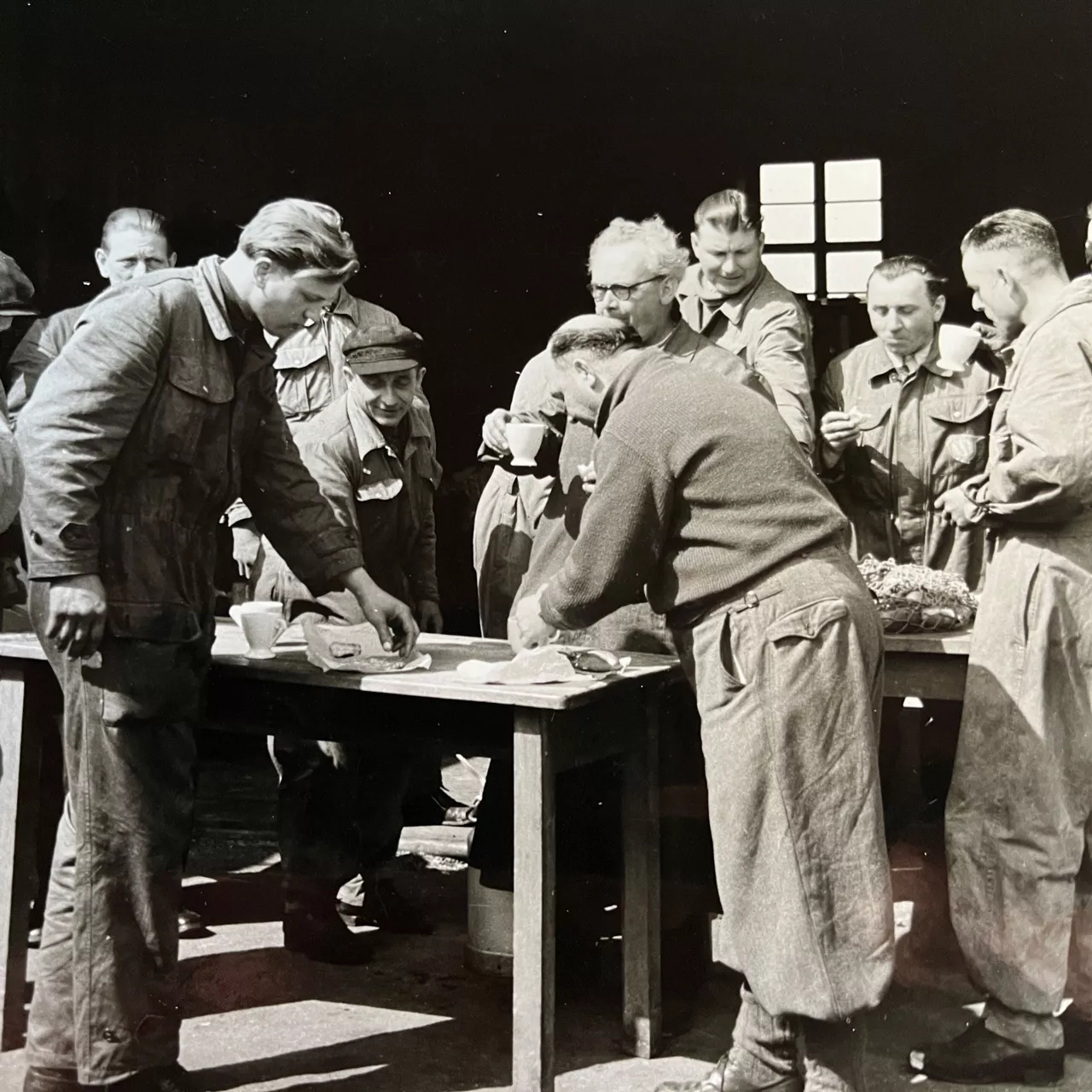 Schwarz-weiß Bild von ein paar Personen stehen um einen Tisch herum und schauen auf Sachen, die auf dem Tisch liegen.