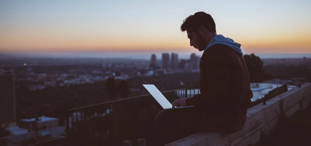 Ein Mann sitzt bei Sonnenaufgang auf einer Mauer und hat seinen Laptop auf den Schoß