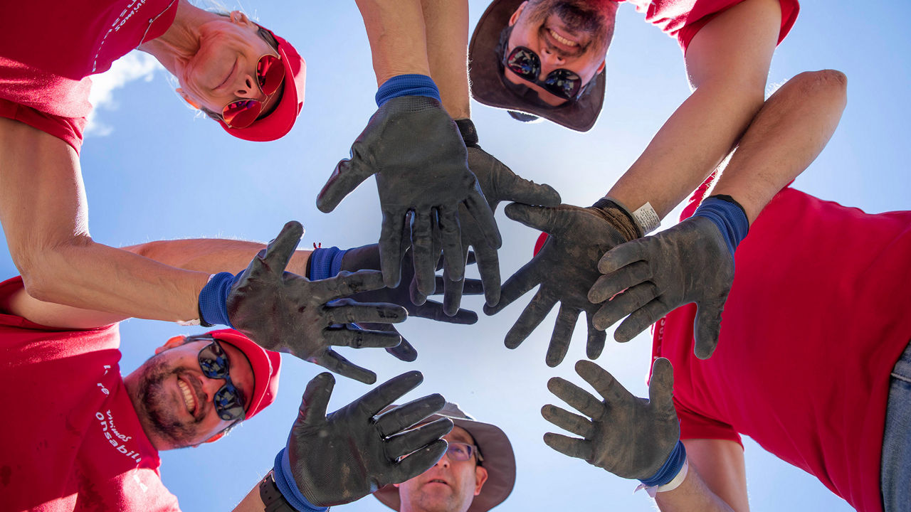 A group of people stand in a circle and stretch their hands towards the center. They are wearing red clothing and look cheerful and motivated as they look up together.