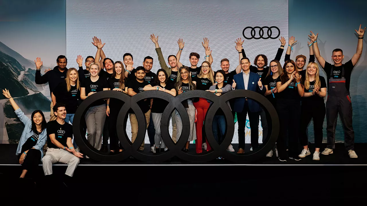 Eine Gruppe Menschen hebt die Arme und steht vor einem großen schwarzen Audi-Logo