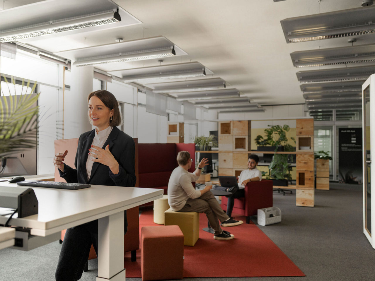Three people are in an office, two are sitting and talking and a woman is standing at a desk talking into a computer screen