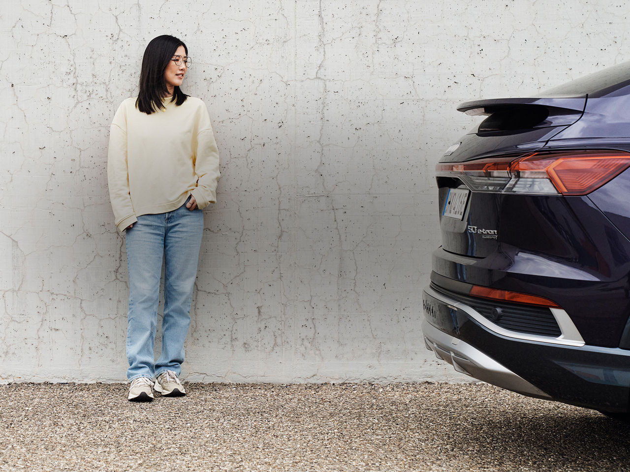 Tiantian stands against a wall and looks to the right at a purple Audi