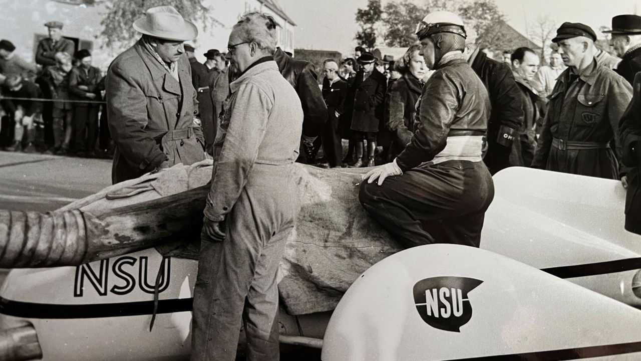 Black and white image of many people standing in front of a vehicle. One person is wearing a helmet
