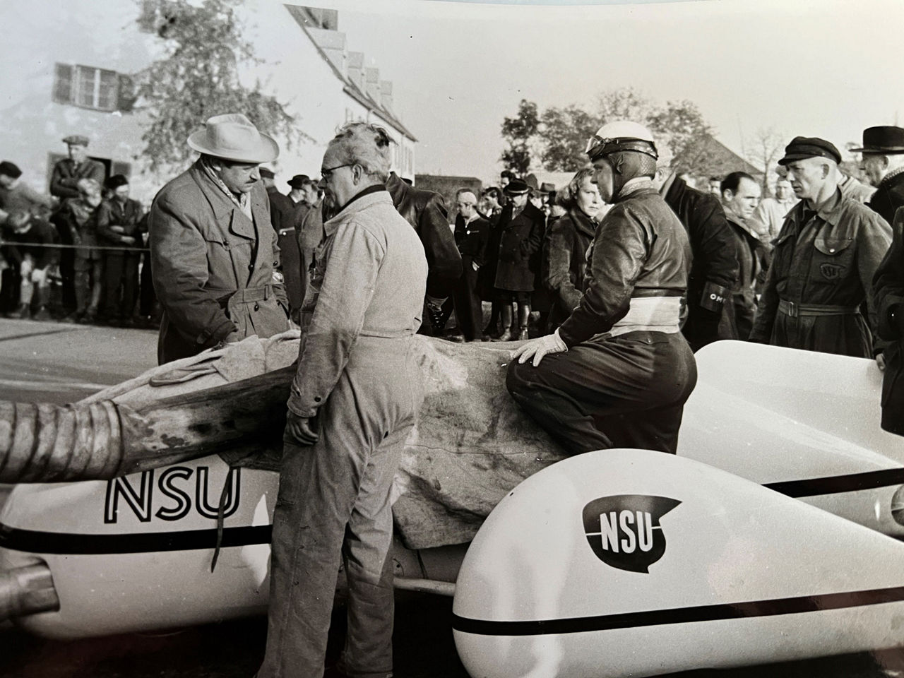 Black and white image of many people standing in front of a vehicle. One person is wearing a helmet