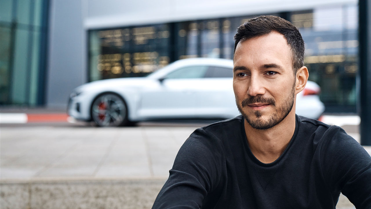 Andreas schaut an der Kamera vorbei und hat einen weißen Audi hinter sich