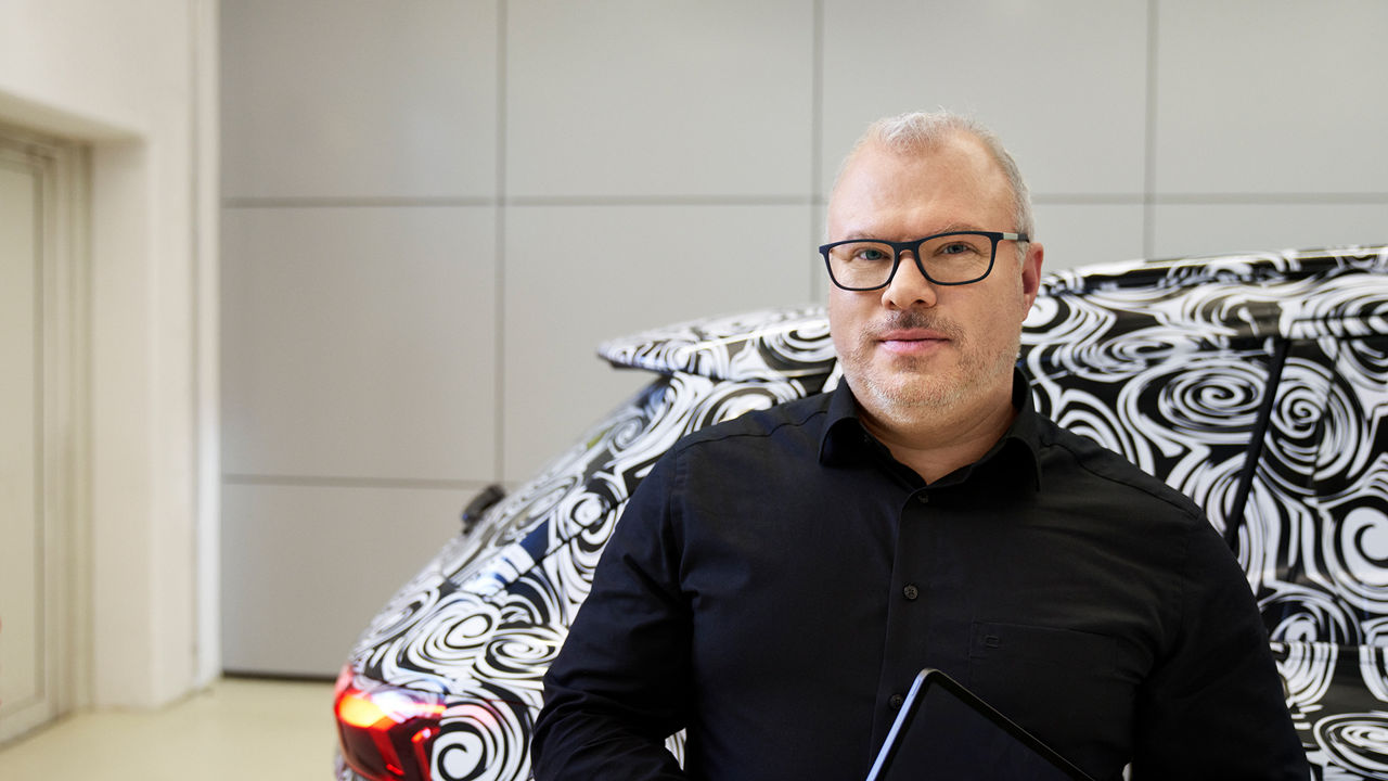 Steffen schaut in die Kamera und hat einen schwarz weißen Audi hinter sich mit einem Muster