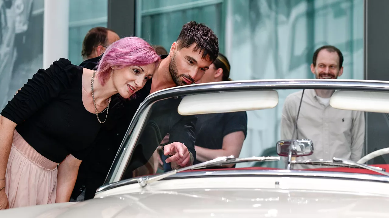 A woman with pink hair and a man look at a classic car together. Other people can be seen in the background.