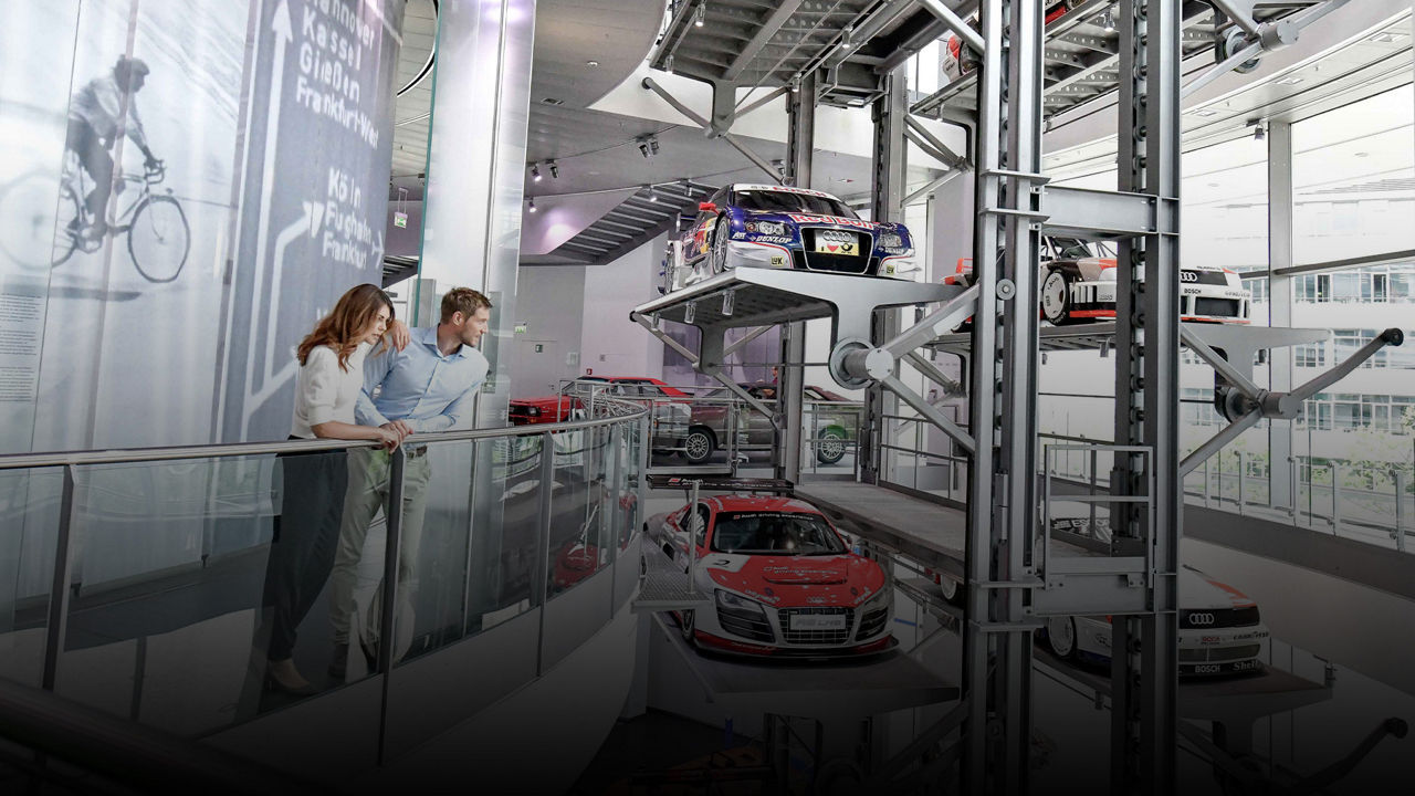 Ein Paar blickt in einem modernen Ausstellungsraum auf rotierende Plattformen mit Renn- und Sportwagen von Audi.