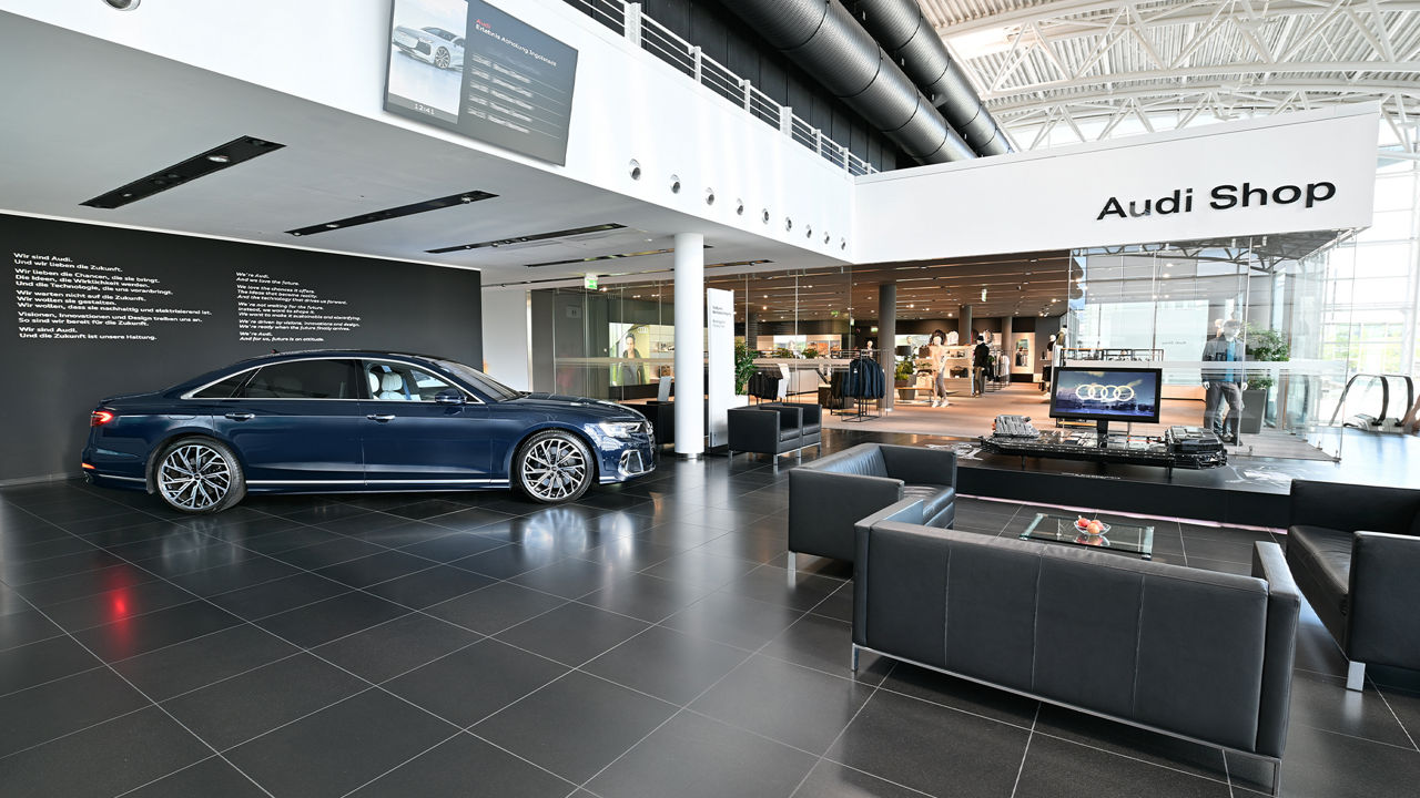 An elegant Audi saloon showroom with a dark blue Audi A8L, surrounded by modern seating areas and an Audi shop in the background.