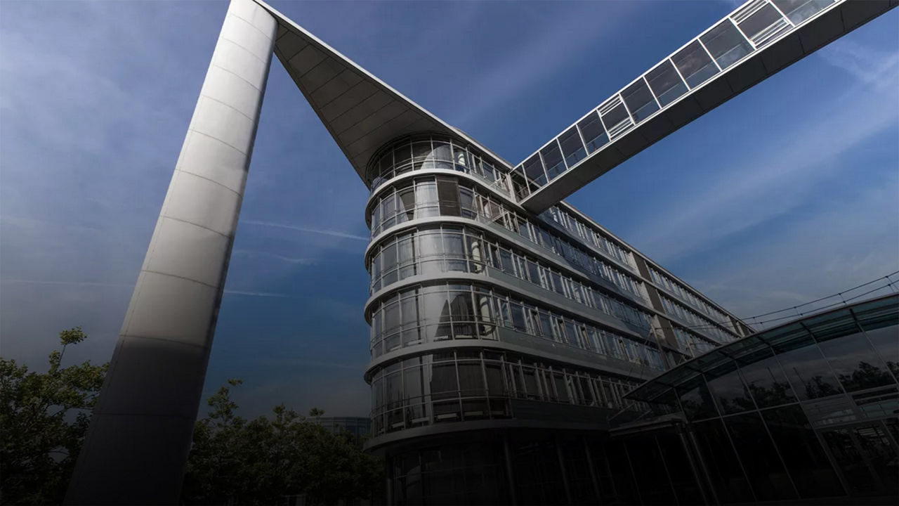 Modernes Bürogebäude mit gläserner Brücke unter blauem Himmel, markante Architektur mit Stützsäule.