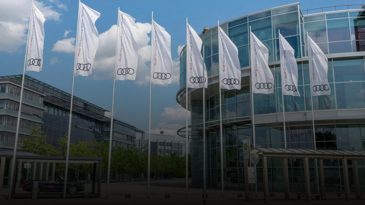Reihe weißer Audi-Fahnen weht vor einem modernen Glasgebäude bei blauem Himmel.