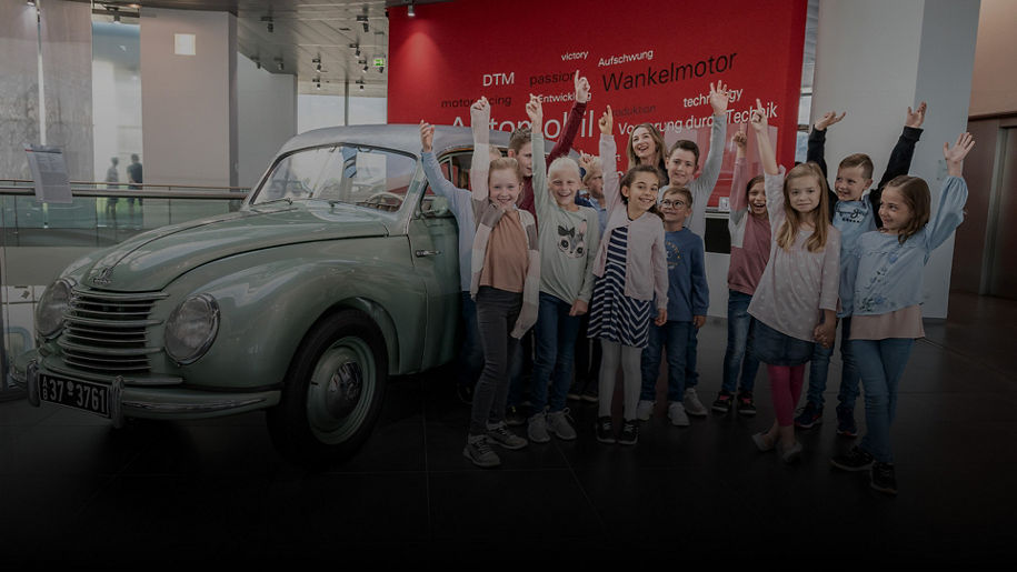 Kindergruppe posiert fröhlich vor einem mintgrünen Oldtimer im Audi Forum; rote Wand mit Technikbegriffen im Hintergrund.