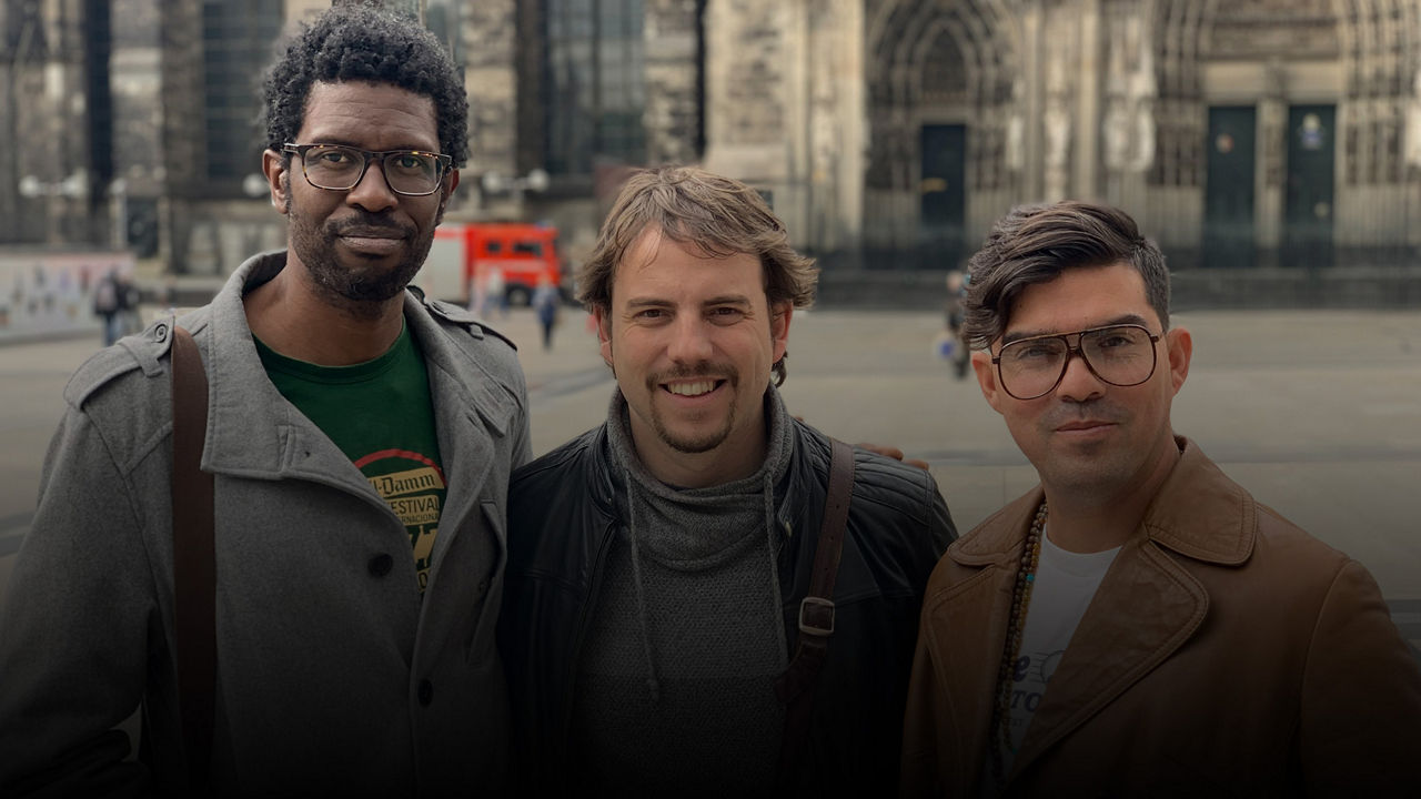 Drei Männer (Daniel Garcia Trio) stehen nebeneinander und blicken lächelnd in die Kamera, im Hintergrund eine große Kirche.