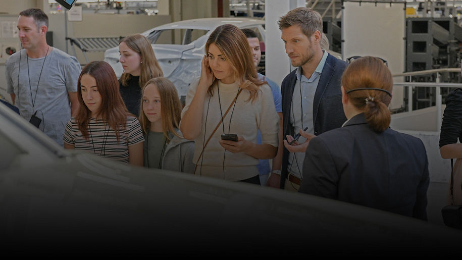 Besuchergruppe mit Audioguide bei Werksführung; Erwachsene und Kinder betrachten interessiert ein Fahrzeug in der Produktion.