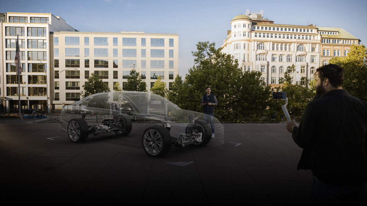Zwei Männer im Freien betrachten ein transparent projiziertes Auto-Modell mit sichtbarer Technik über einem Stadtplatz.