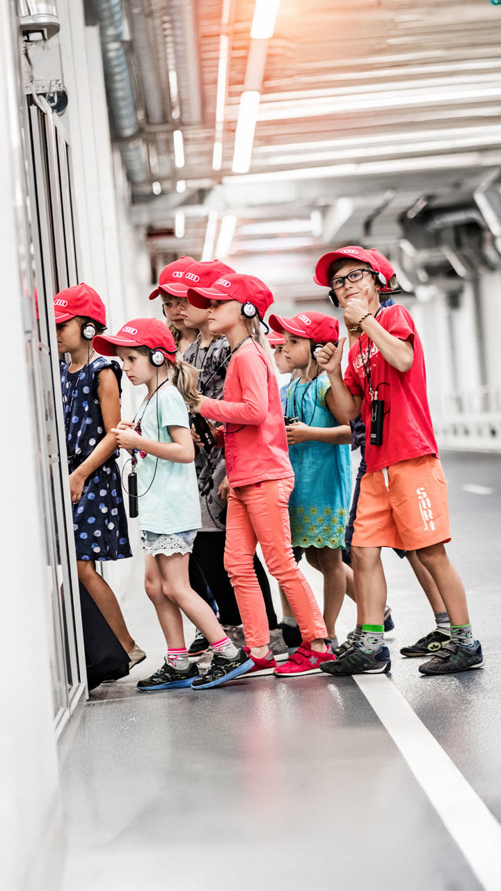 Eine Gruppe von sechs Kindern mit roten Kappen und Kopfhörern steht nebeneinander und schaut aufmerksam in eine Ausstellungsraum.