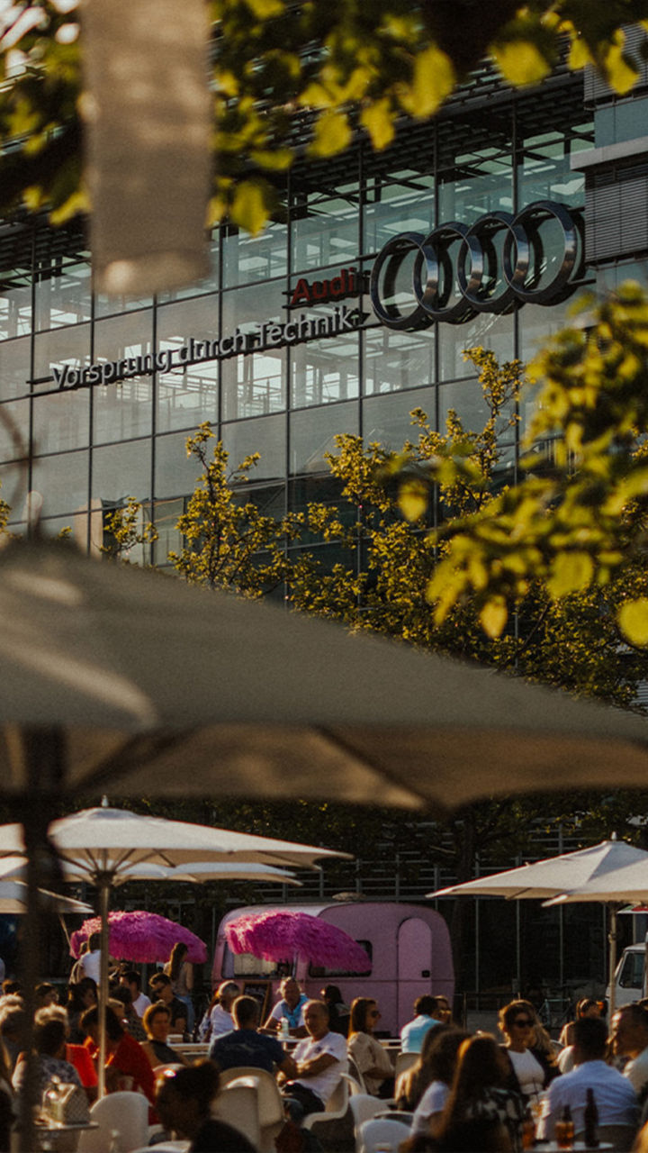 Menschen sitzen unter Sonnenschirmen bei sommerlichem Wetter vor dem Audi-Gebäude mit „Vorsprung durch Technik“-Schriftzug.