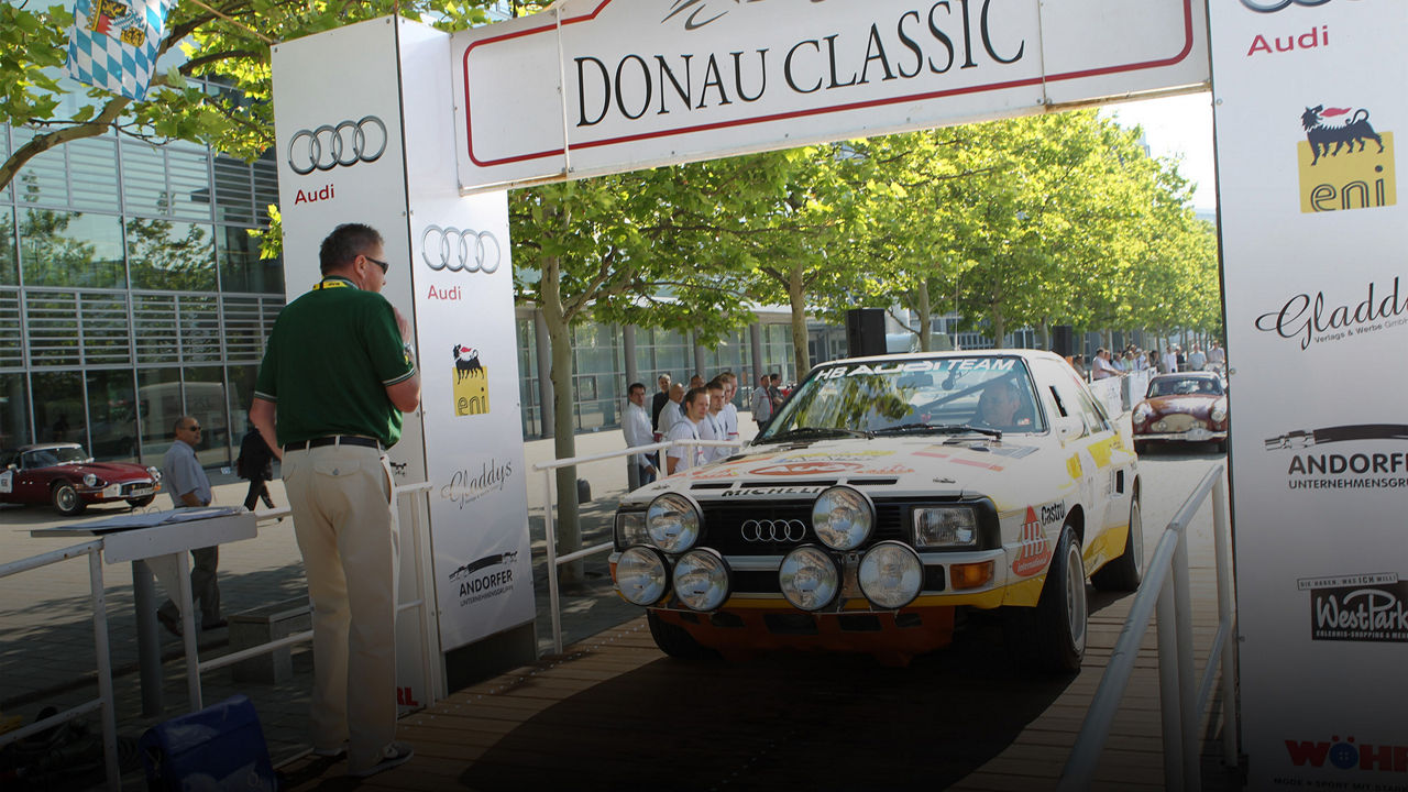 Ein weißer Audi Rallye Quattro mit gelben Akzenten fährt durch einen Torbogen der "DONAU CLASSIC". Ein Mann in grünem Shirt beobachtet das Auto.