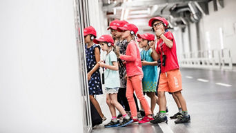 Sechs Kinder mit roten Kappen und Kopfhörern betrachten eine Ausstellungsvitrine. 