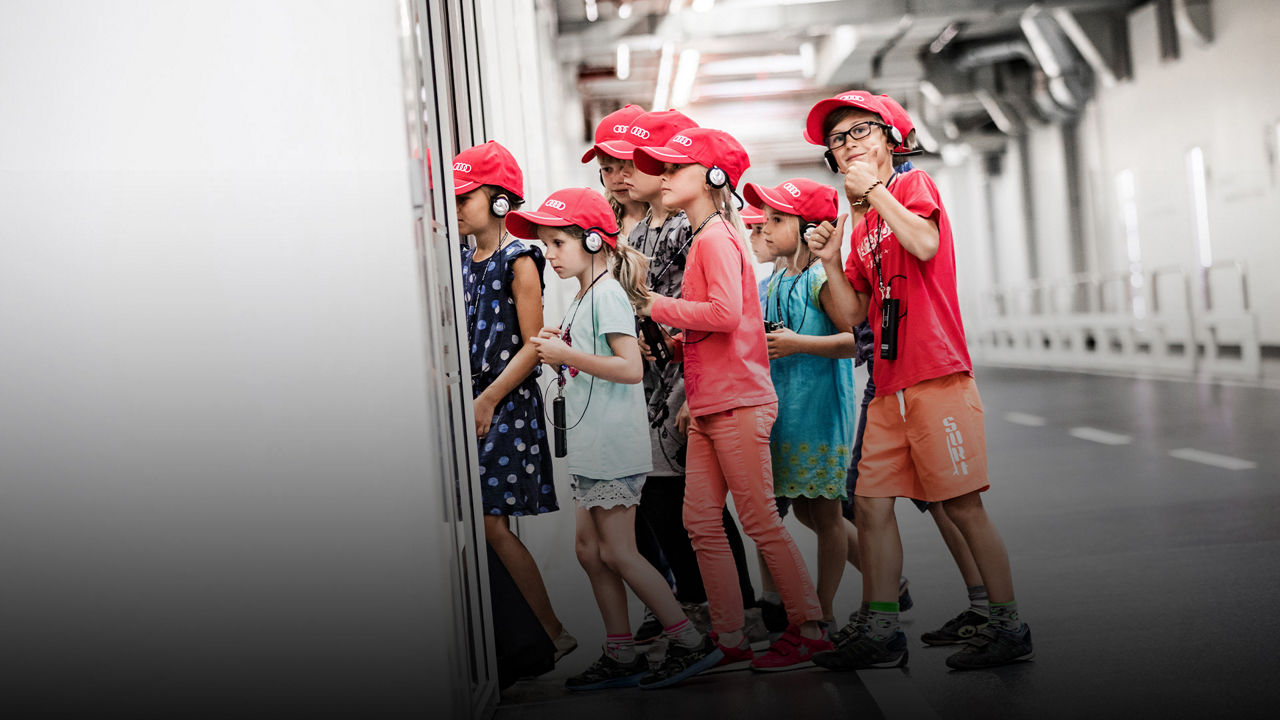  Eine Gruppe von sechs Kindern mit roten Kappen und Kopfhörern steht nebeneinander und schaut aufmerksam in eine Ausstellungsraum. 