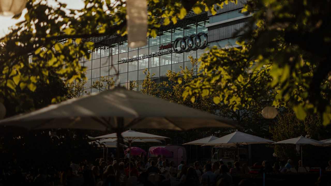  Eine sonnige Außenansicht mit grünen Bäumen und dem Audi-Logo auf einem modernen Gebäude im Hintergrund. Im Vordergrund sind Menschen auf einer Terrasse unter Sonnenschirmen zu sehen, mit einem rosafarbenen Imbisswagen in der Mitte.