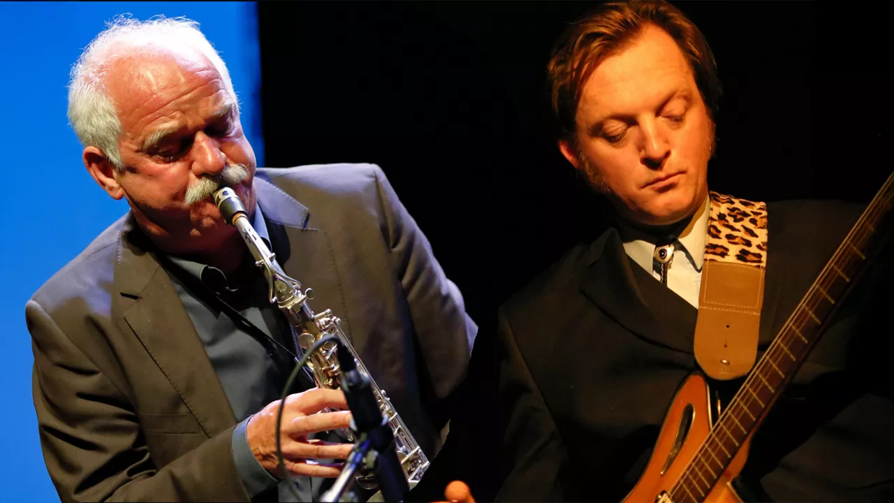 Zwei Musiker spielen Saxophon und Gitarre auf einer Bühne mit dunklem Hintergrund und Bühnenlicht.