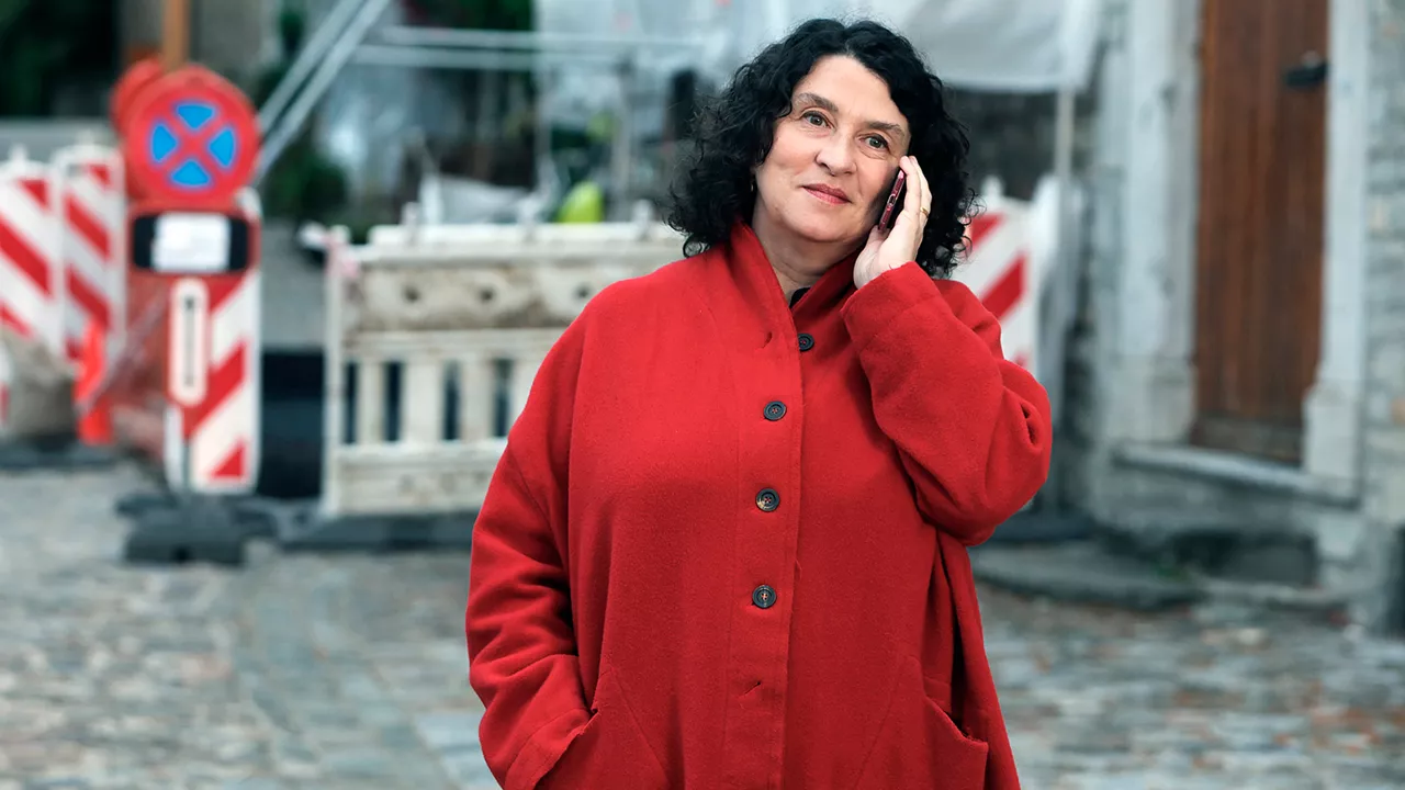 Ältere Frau mit dunklen Locken und einem roten Mantel steht draußen vor einer Baustelle und telefoniert