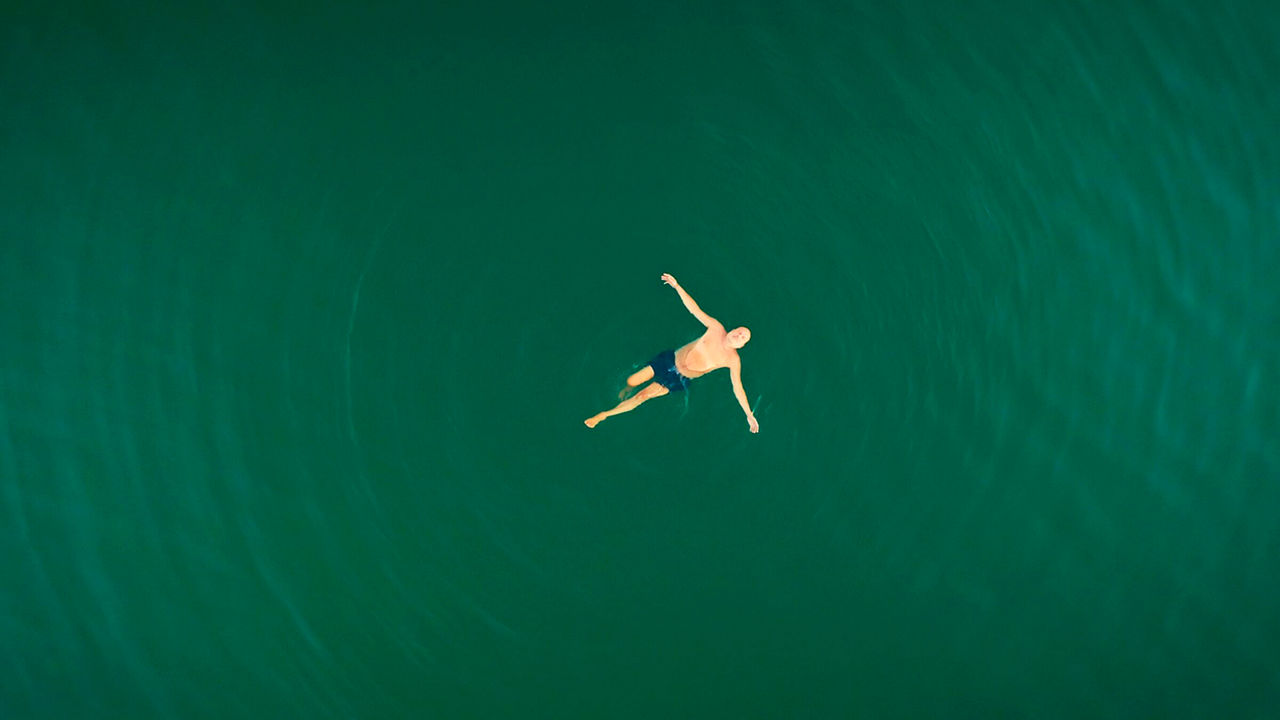 Ein Mann in Badehose liegt in einem Meer mit ausgebreiteten Armen und schaut in den Himmel