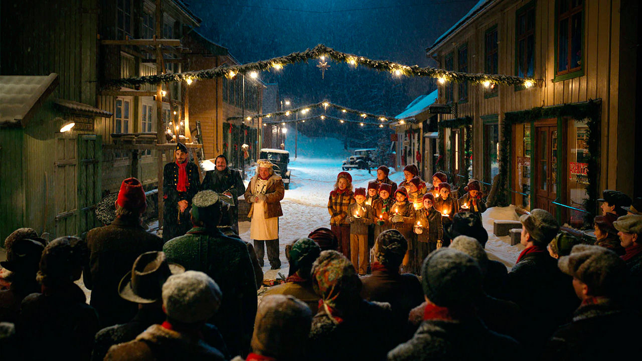 Menschen stehen draußen im Schnee zwischen weihnachtlich geschmückten Häuserreihen, Gruppe von Kindern halten eine Kerze in der Hand 