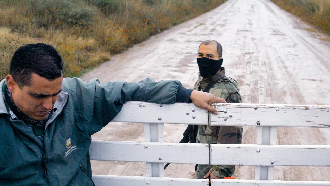 2 Männer sind auf einer kaputten Straße in der Natur. Der eine lehnt an einem weißen Zaun und der andere trägt eine schwarze Maske am Mund und steht mit einem Gewähr