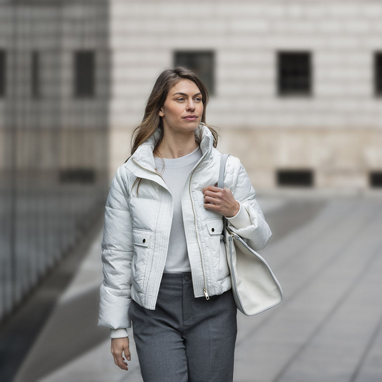 Eine Frau mit einer weissen Jacke beleidet geht an einem Gebäude mit einer verspiegelten Glasfront vorbei. Über die Schulter trägt sie eine weisse Handtasche.