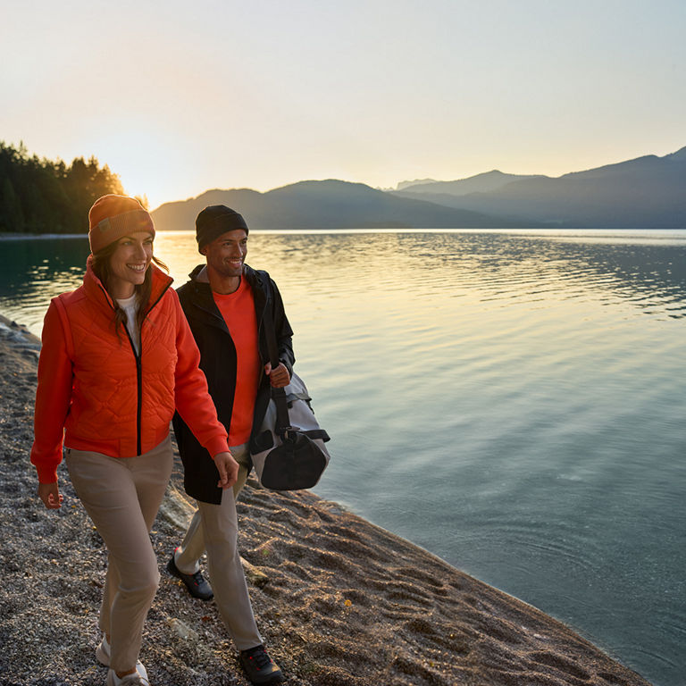 Eine Mann und eine Frau spazieren bei Sonnenuntergang an einem See entlang. Er trägt über die Schulter eine graue Audi Sporttasche.