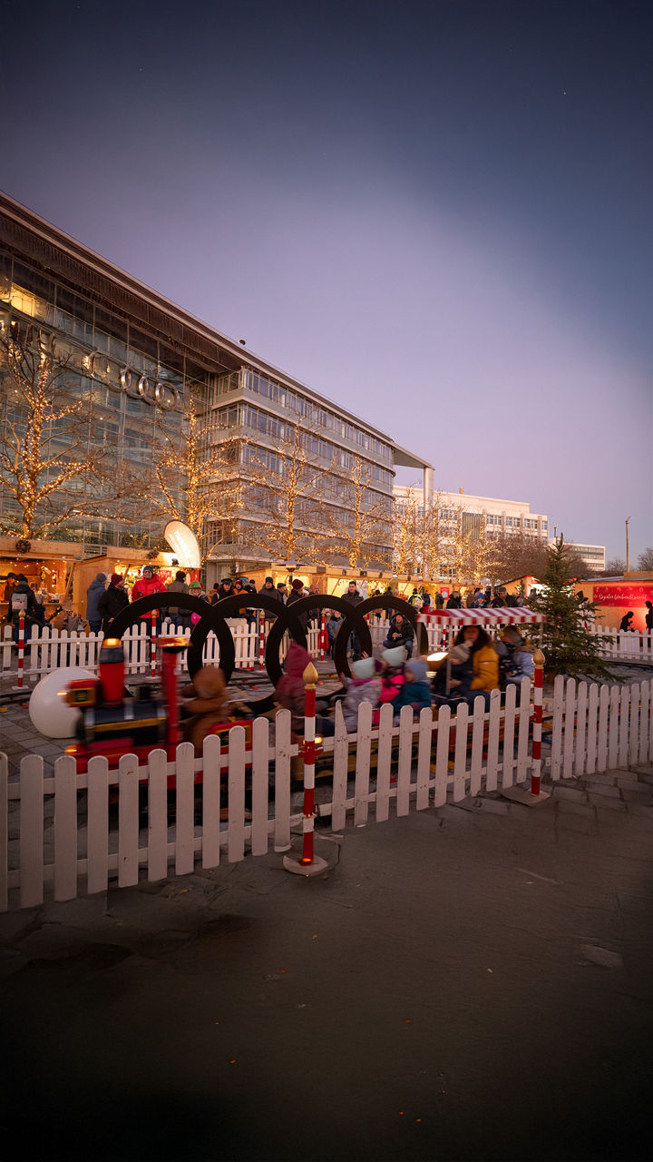 Kinder fahren in einer kleinen Bahn auf einem Weihnachtsmarkt; im Hintergrund Menschen, Buden und beleuchtete Bäume.