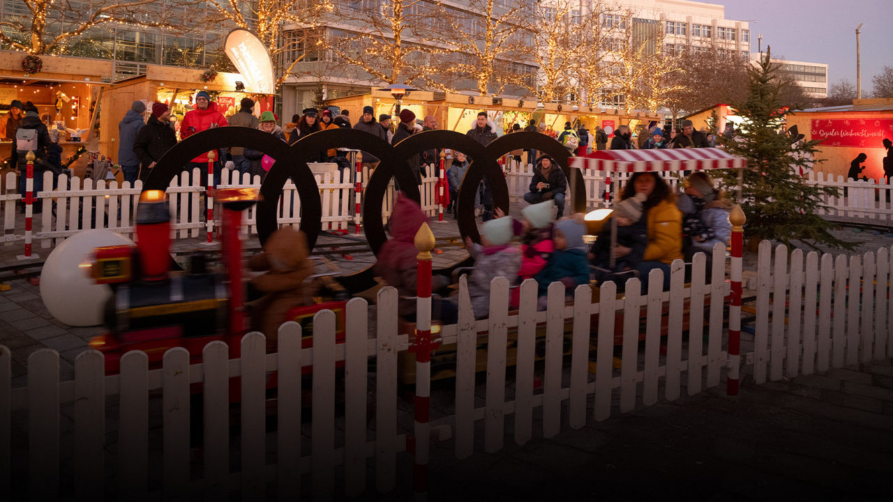 Kinder fahren in einer kleinen Bahn auf einem Weihnachtsmarkt; im Hintergrund Menschen, Buden und beleuchtete Bäume.