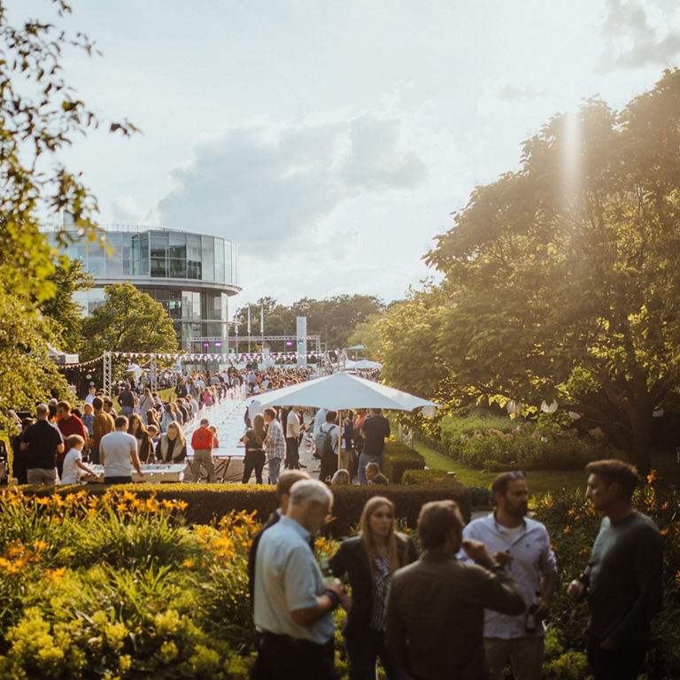 Menschen stehen in einer parkähnlichen Anlage. Im Hintergrund ist das Audi museum mobile zu sehen.