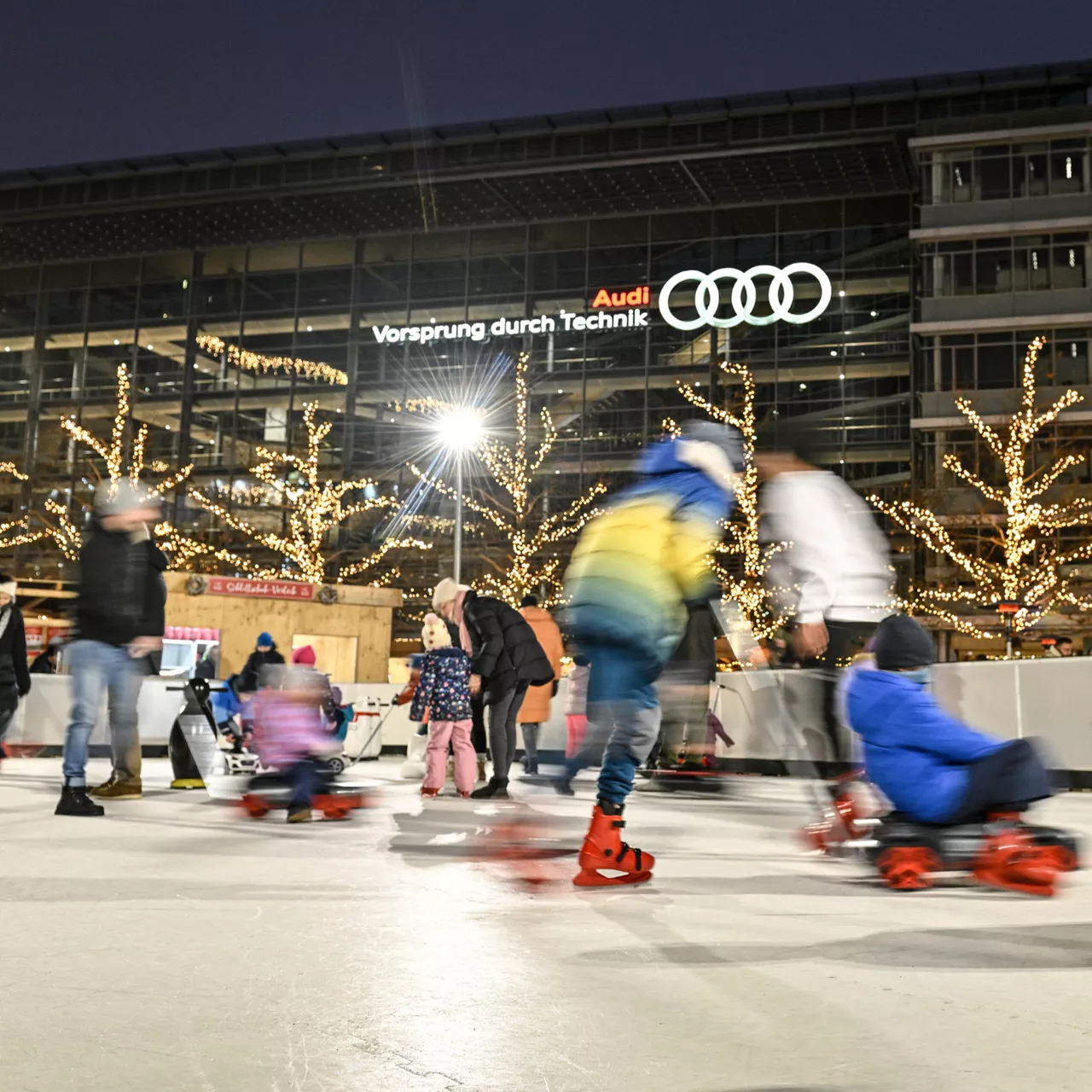 Kinder und Erwachsene fahren Schlittschuh auf einer Eisbahn; im Hintergrund Audi-Gebäude und beleuchtete Bäume.