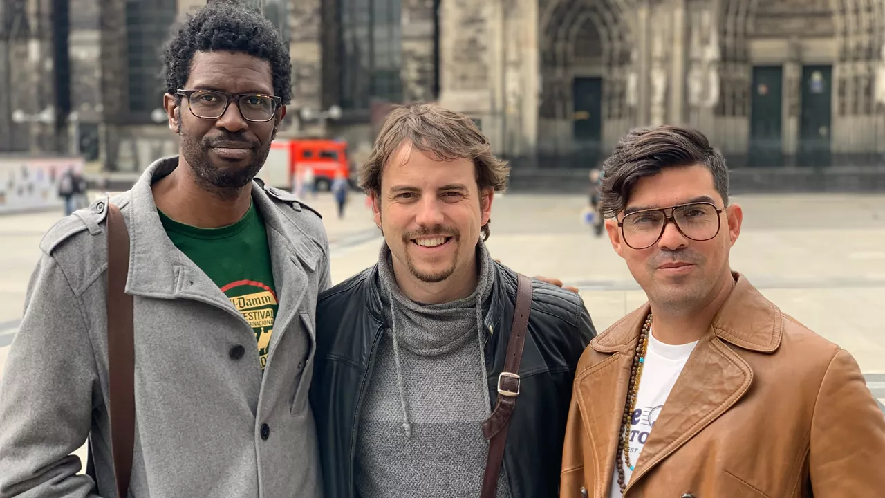 Drei Männer (Daniel Garcia Trio) stehen nebeneinander und blicken lächelnd in die Kamera, im Hintergrund eine große Kirche.