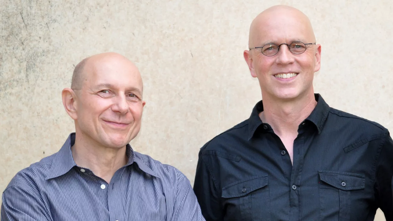 Zwei Männer lächeln in die Kamera, beide tragen Hemden, einer davon eine Brille.