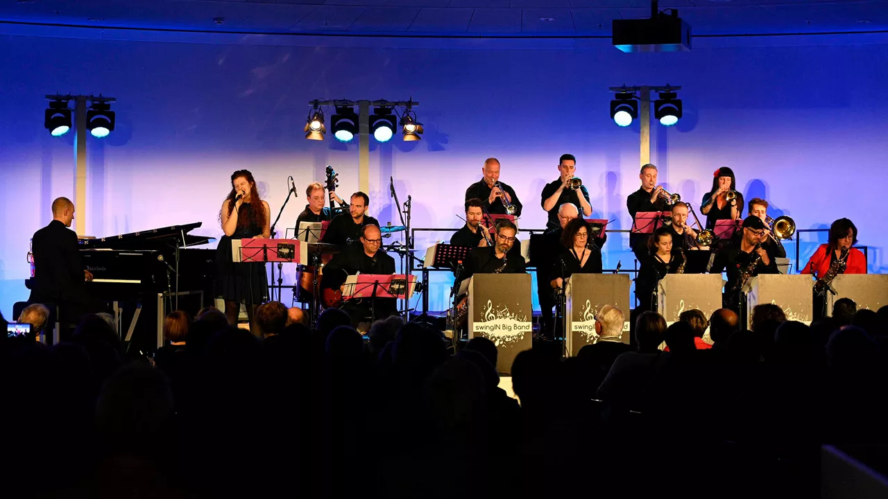 Große Bigband mit Sängerin (SwingIN Big Band) bei Konzertauftritt auf einer Bühne vor Publikum, beleuchtet in Blau und Violett.