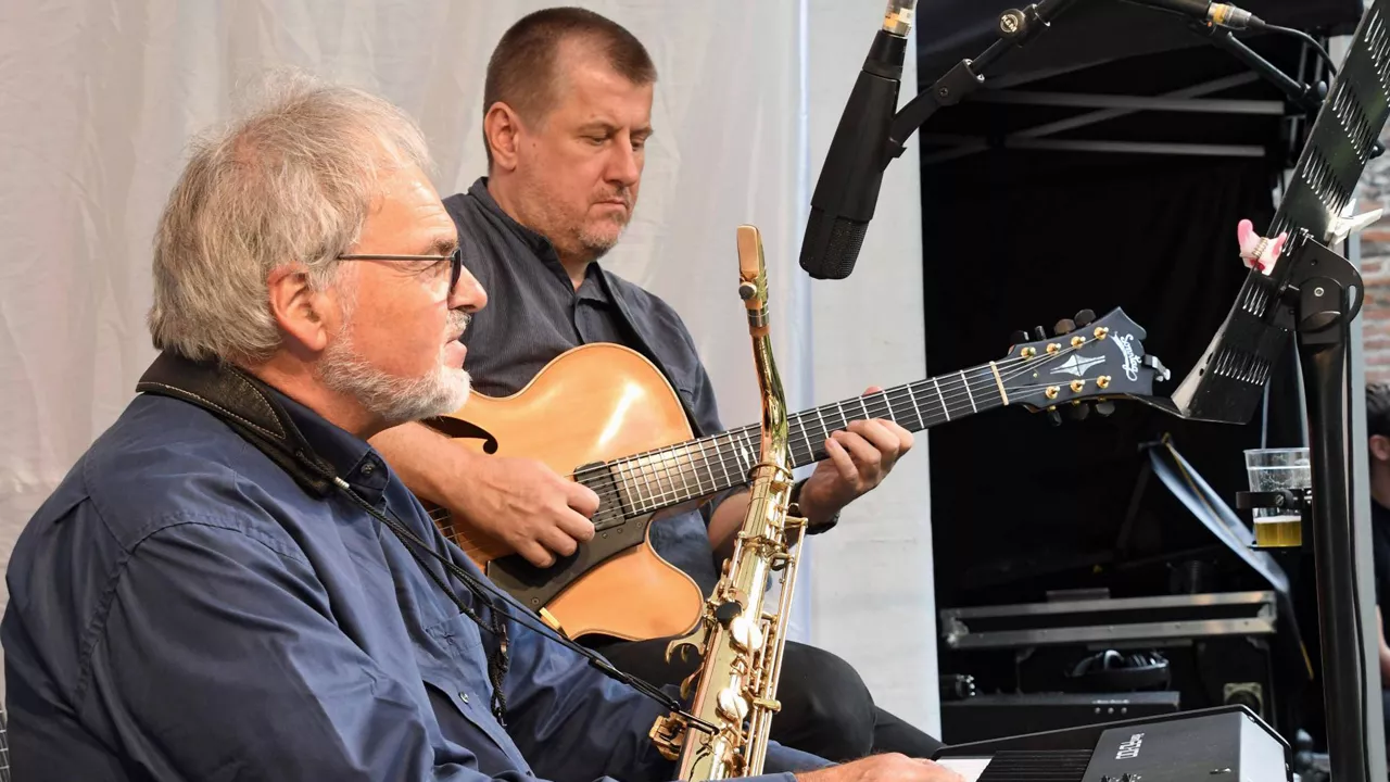 Zwei Musiker, einer spielt Saxophon, der andere Gitarre, vor Mikrofonen auf einer Bühne.
