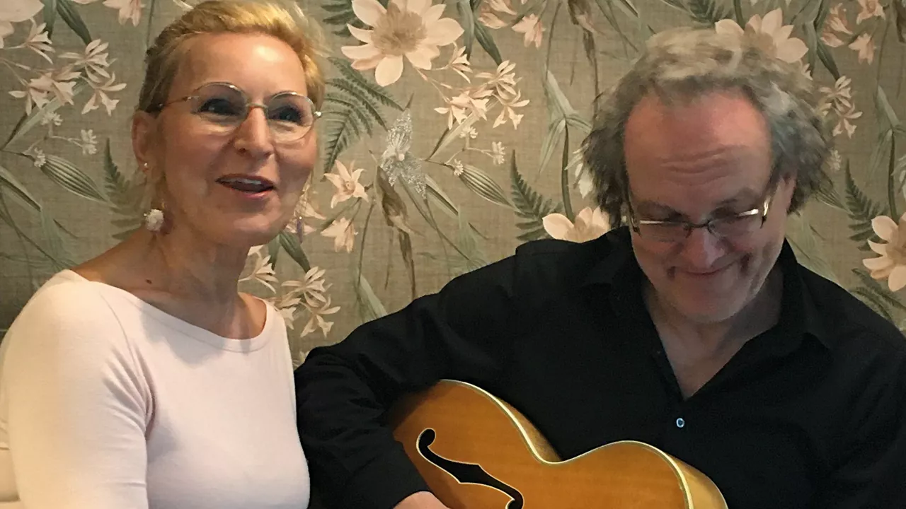 Frau singt neben einem Mann, der eine Gitarre spielt, vor blumiger Tapete.