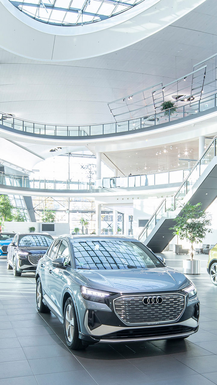 A spacious, light-flooded showroom with several Audi models, including a silver electric SUV and a golden Q5, as well as modern architecture with glass railings and a curved staircase.