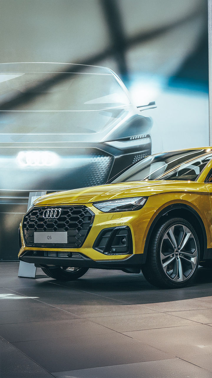 A golden yellow Audi Q5 stands in a showroom in front of a large wall showing a futuristic vehicle study with a glowing Audi logo.