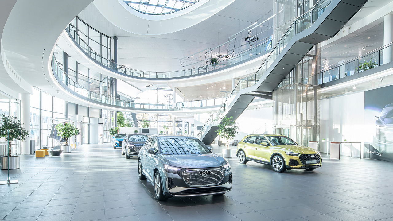 A spacious, light-flooded showroom with several Audi models, including a silver electric SUV and a golden Q5, as well as modern architecture with glass railings and a curved staircase.