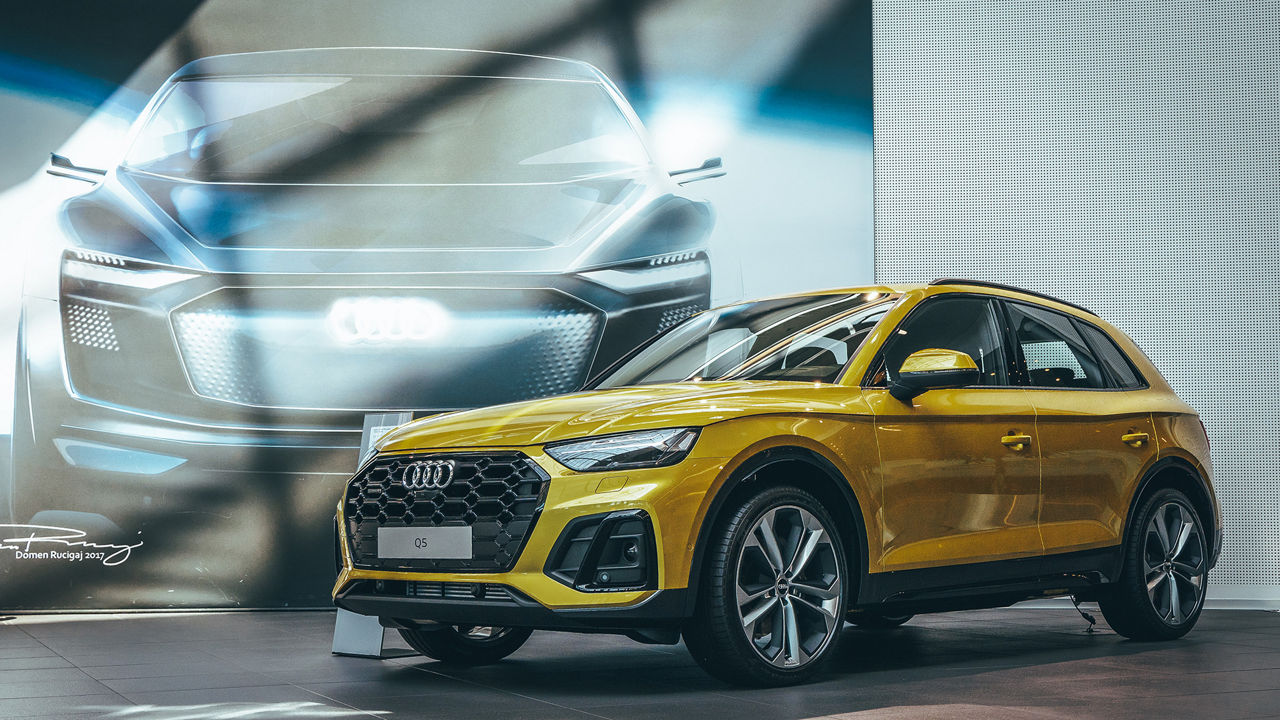 A golden yellow Audi Q5 stands in a showroom in front of a large wall showing a futuristic vehicle study with a glowing Audi logo.