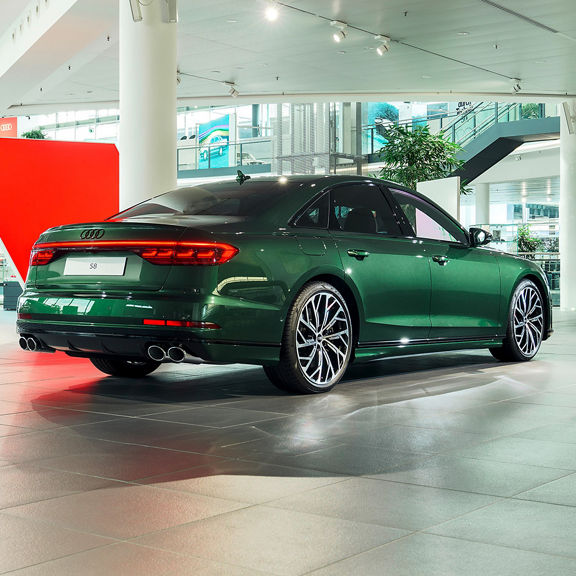 A dark green Audi S8 stands in a modern, light-flooded showroom with large windows and stylish architecture.