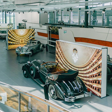 A showroom with several classic cars, including a black cabriolet in the foreground. The vehicles are presented in front of artistic backdrops with opera or theatre hall motifs.